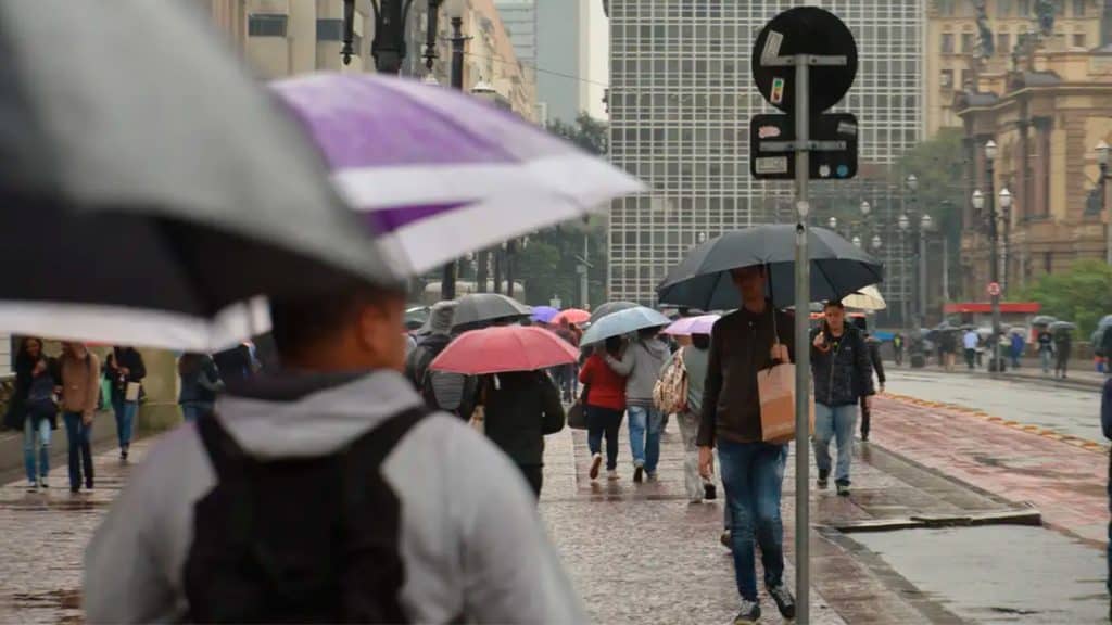 Pessoas andando em calçada carregando guarda-chuvas em São Paulo